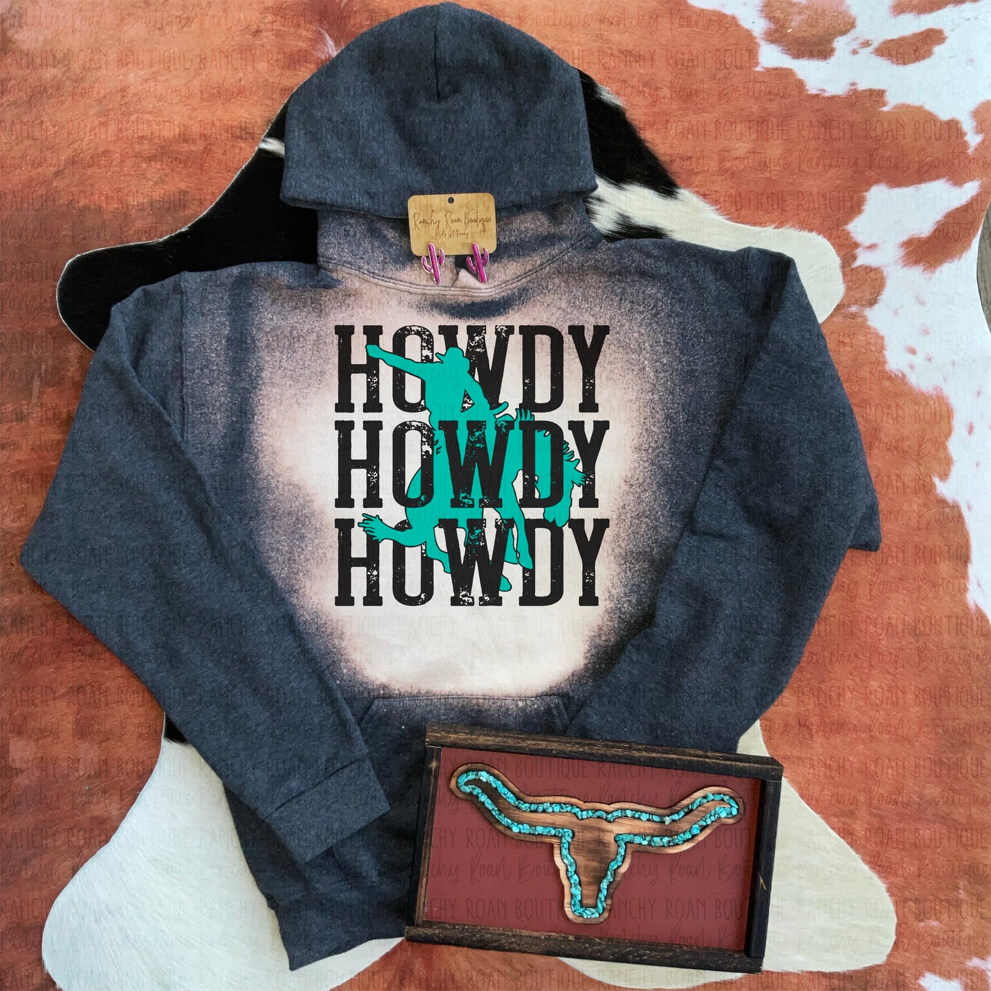 Heather Black  bleached “Howdy” graphic hoodie with turquoise bronc rider silhouette, displayed with turquoise earrings and decorative skull on a cowhide background.
