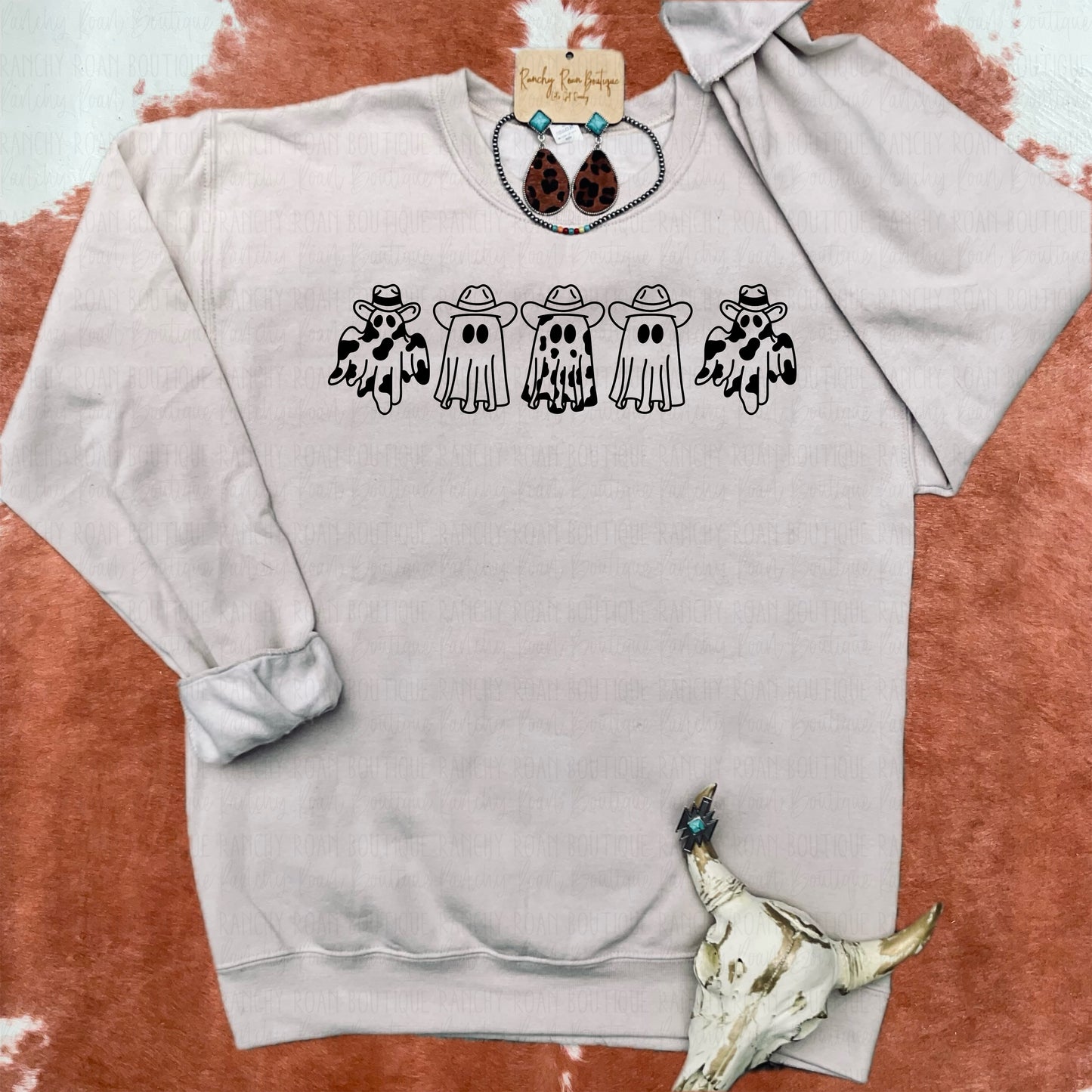 A sandstone color sweatshirt from Ranchy Roan Boutique featuring a row of playful ghosts dressed in Western attire, including cowboy hats and bandanas, giving it a fun and festive look.