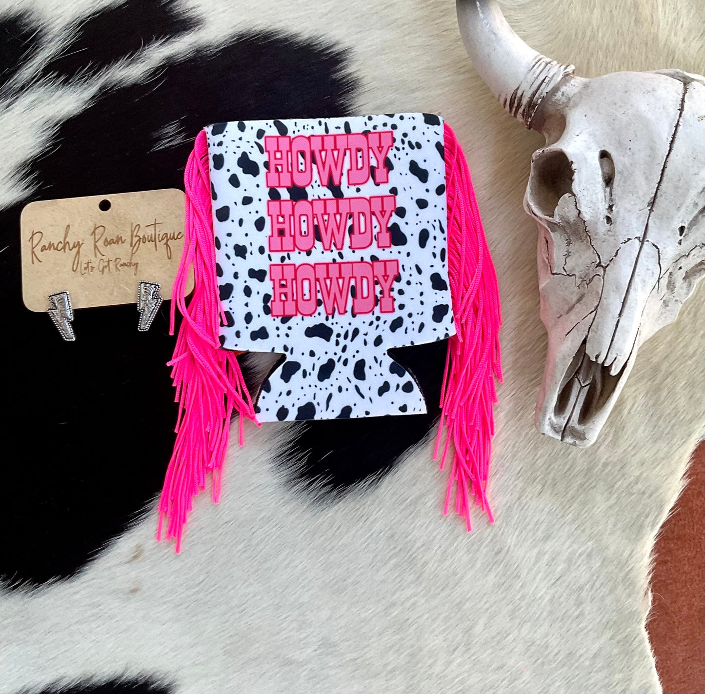 ‘Howdy Howdy Howdy” cow print koozie with hot pink fringe, displayed on a cowhide background with Western decor.