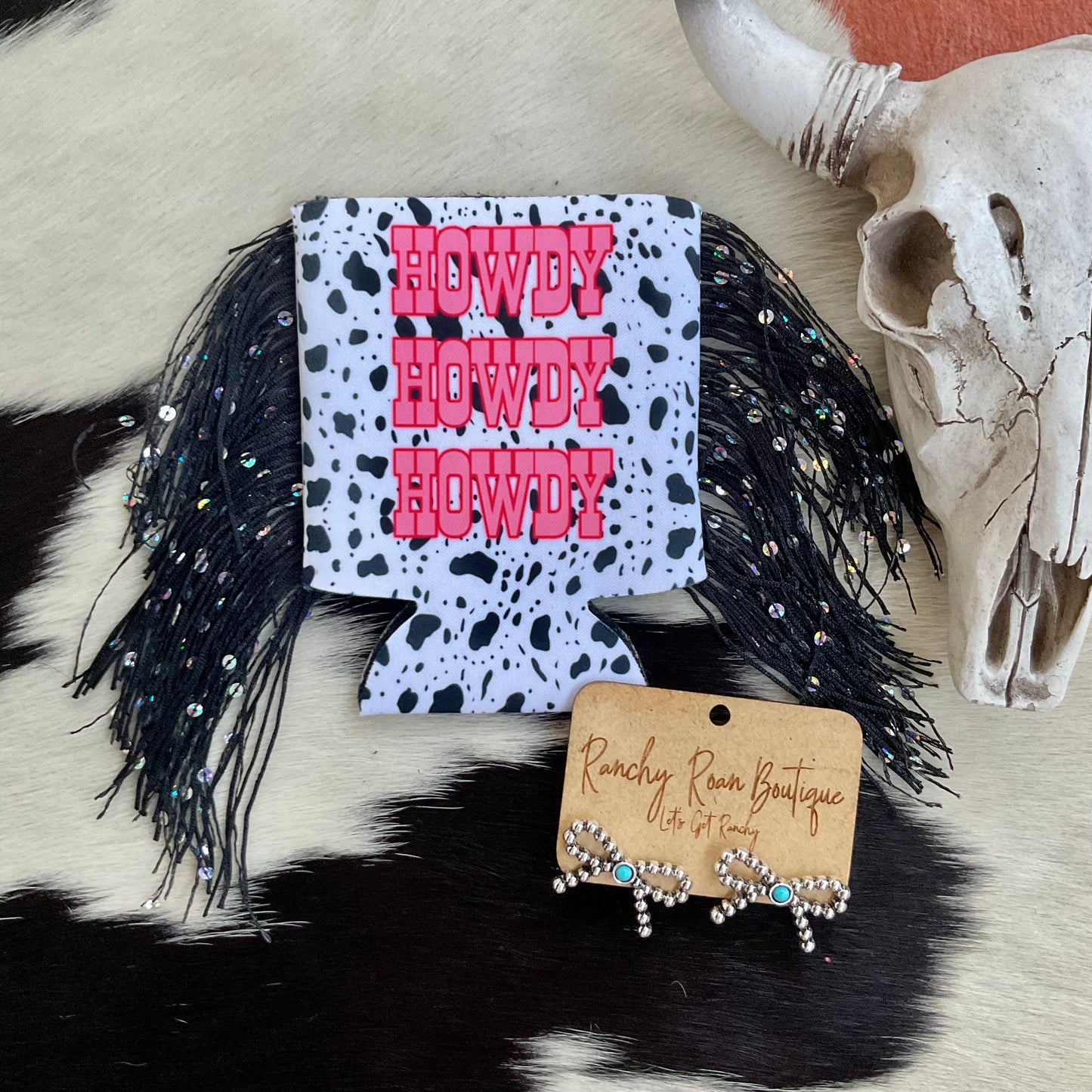 Cow print koozie with bold pink “Howdy” text and black fringe, styled on a Western background.