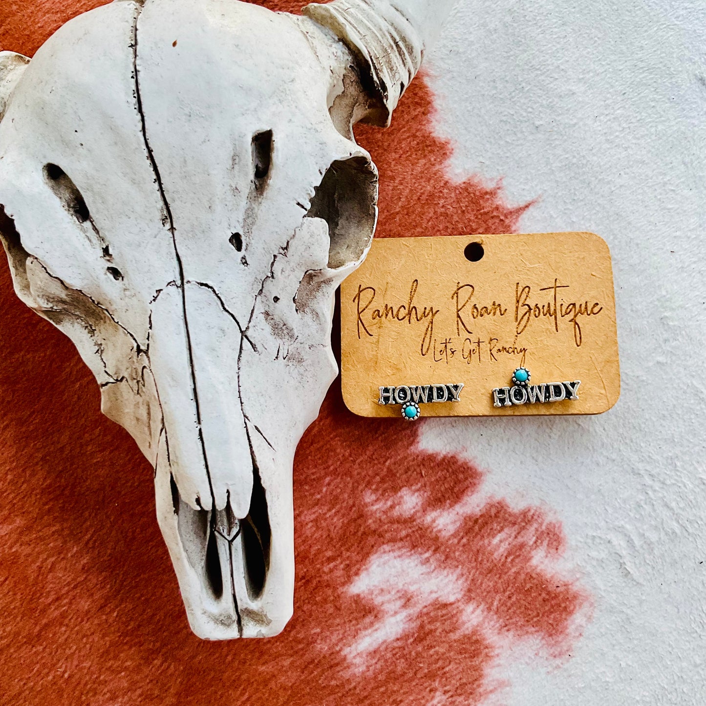 A pair of “HOWDY” word earrings with turquoise accents displayed on a Ranchy Roan Boutique card against a cowhide background, accompanied by a decorative cow skull.