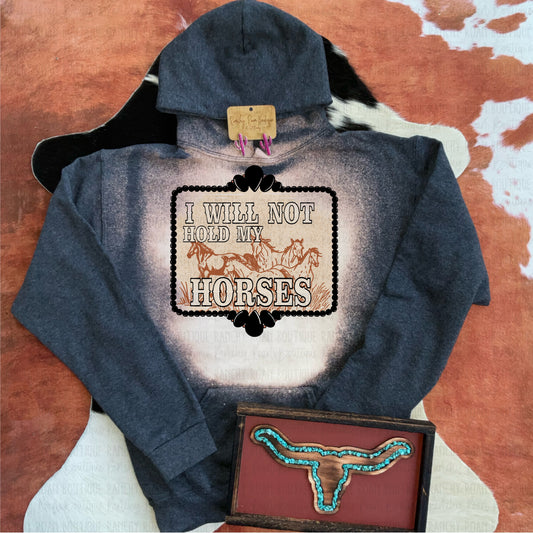 A bleached hoodie from Ranchy Roan Boutique featuring a bold sublimation print with the text “I Will Not Hold My Horses” and a dynamic horse graphic. The hoodie has a unique bleached pattern in shades of dark blue and cream, creating a one-of-a-kind look. Styled with a pair of pink earrings and a turquoise inlay longhorn wall decor, enhancing the Western aesthetic. The background includes a cowhide texture, further emphasizing the rustic, Western theme.