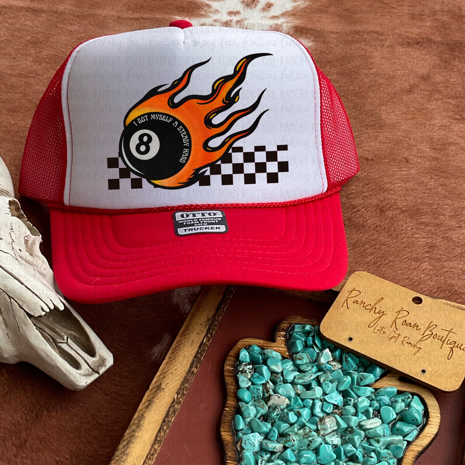 Bold red trucker hat with a flaming 8-ball and checkered graphic displayed on a cowhide background, paired with a Western skull prop.