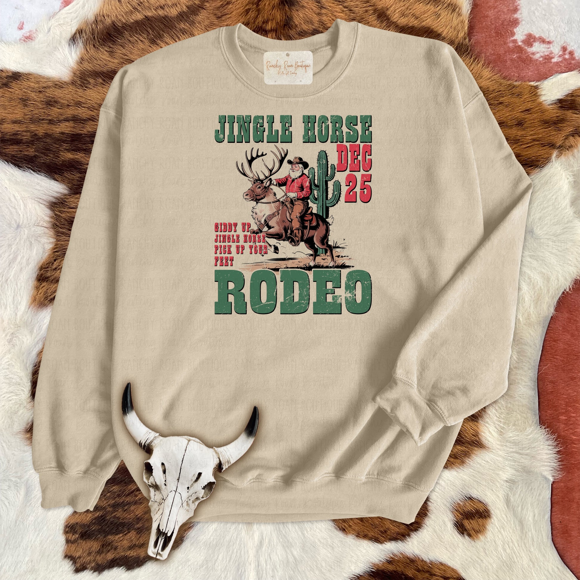 Tan sweatshirt featuring a festive Western design with a cowboy riding a reindeer and the text “Jingle Horse Dec 25 Rodeo.”