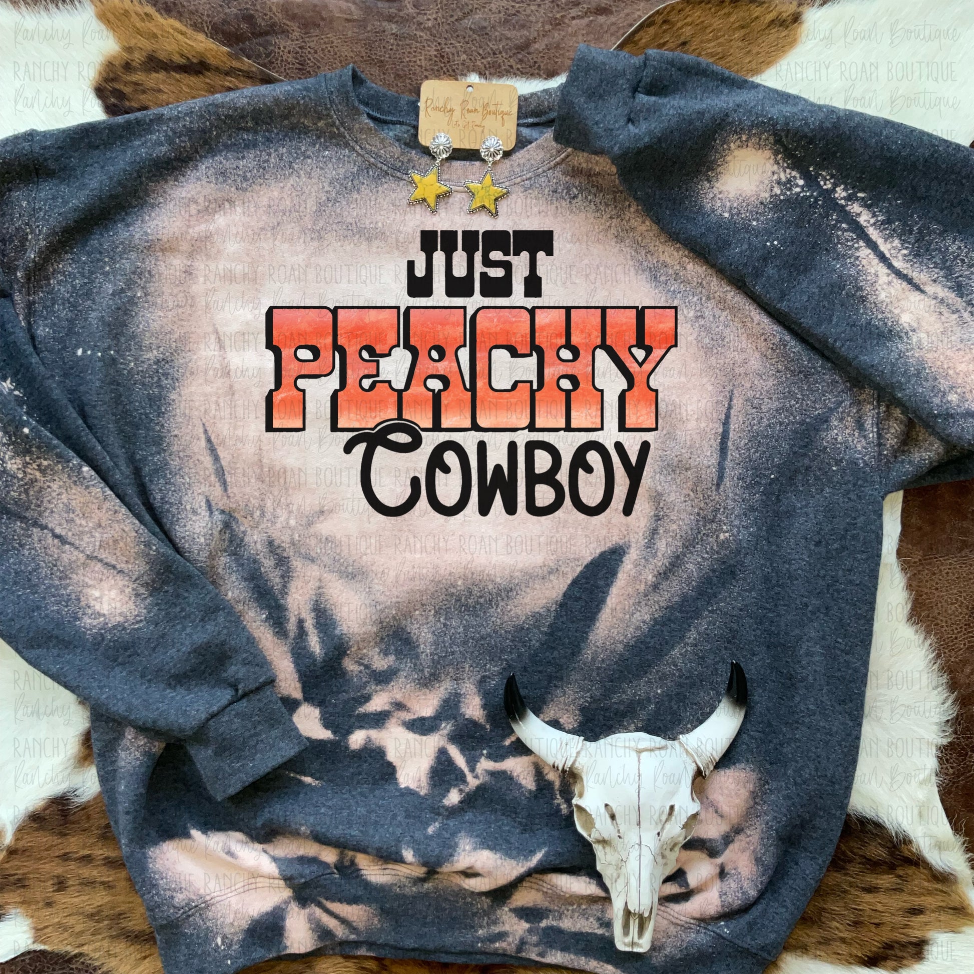 Just Peachy Cowboy” Western-themed bleached sweatshirt, featuring a distressed design with a bold peachy-orange text graphic, styled with star earrings on a rustic background.