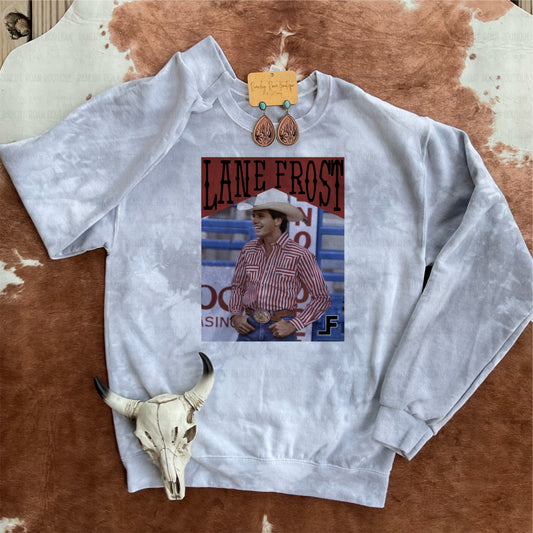 Graphic sweatshirt featuring Lane Frost in a red-striped shirt, styled on a western-themed backdrop with leather earrings and a cow skull accent.