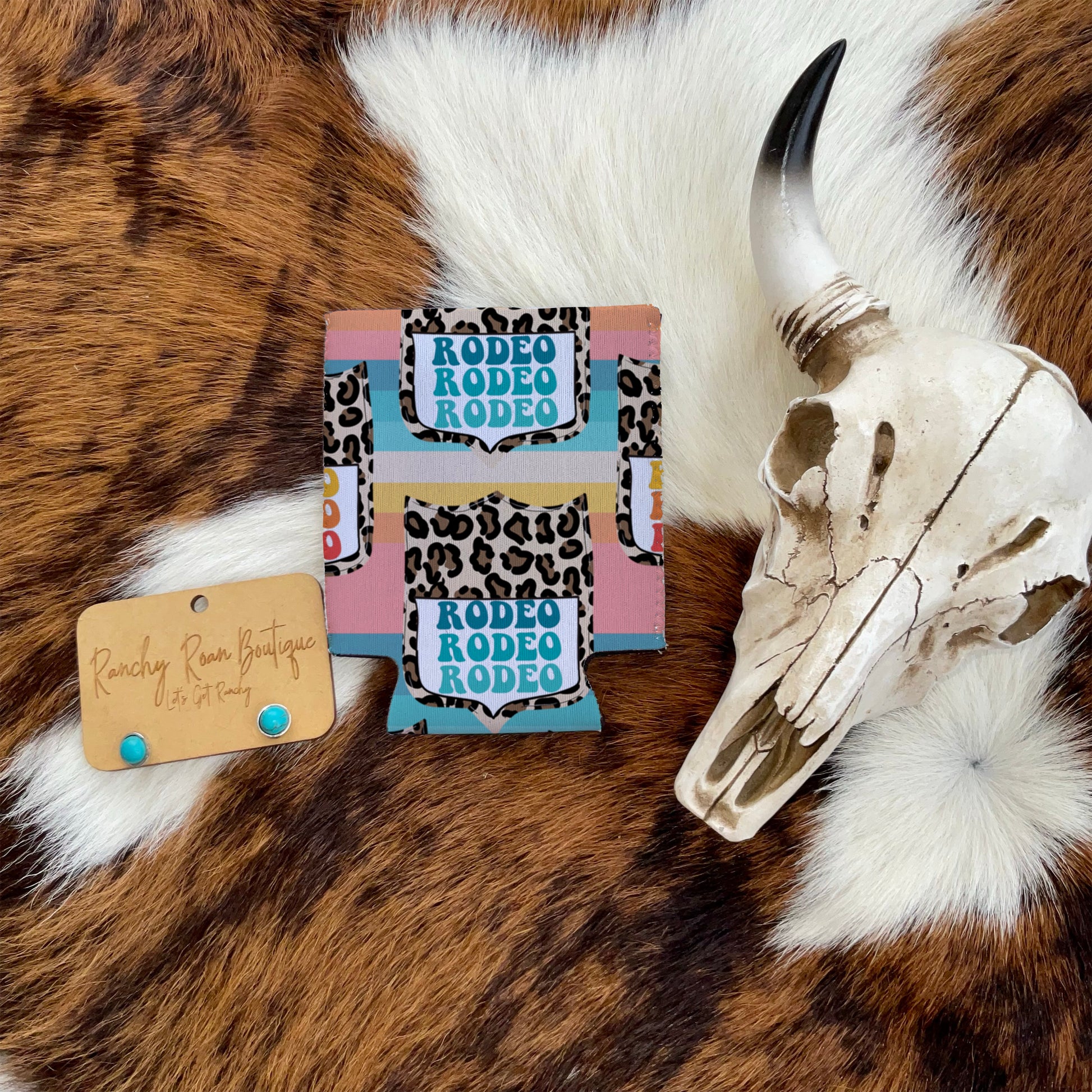 Leopard Rodeo Koozie with bold “Rodeo” text displayed alongside turquoise stud earrings and a decorative cow skull.
