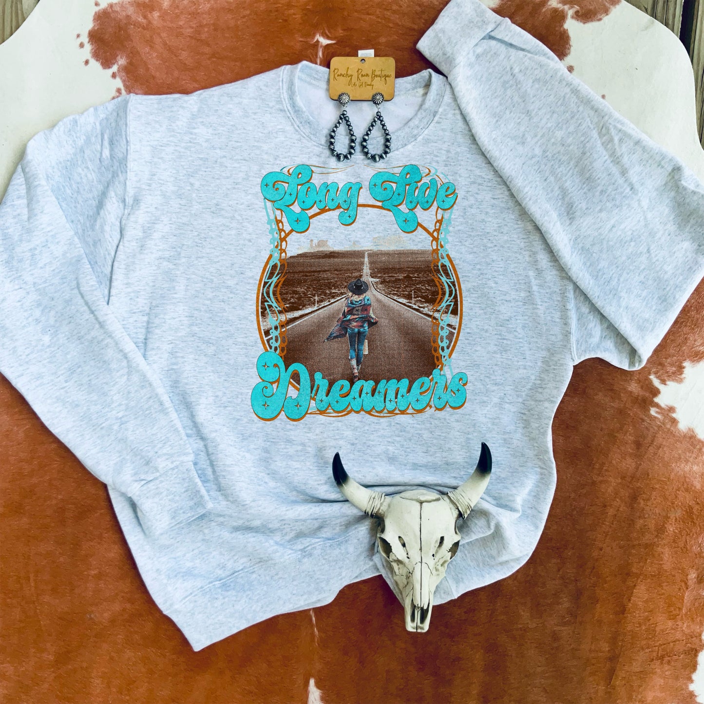 Long Live Dreamers sweatshirt laid flat on a cowhide backdrop, showcasing its Western-inspired graphic and vibrant turquoise text.