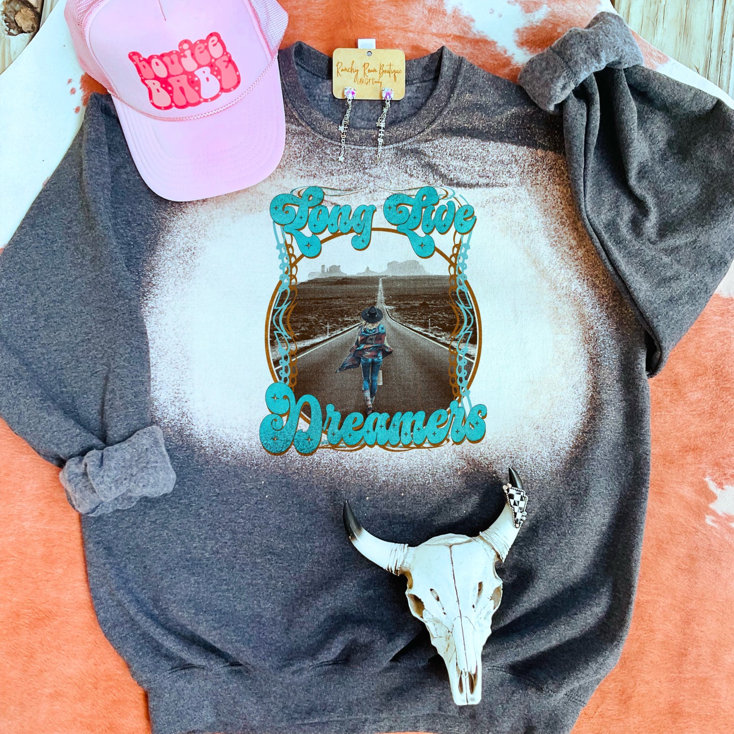 A dark heather-gray unisex sweatshirt with a bleach splatter design and a Western graphic that reads “Long Live Dreamers.” Displayed with a pink trucker hat and Western-themed accessories.
