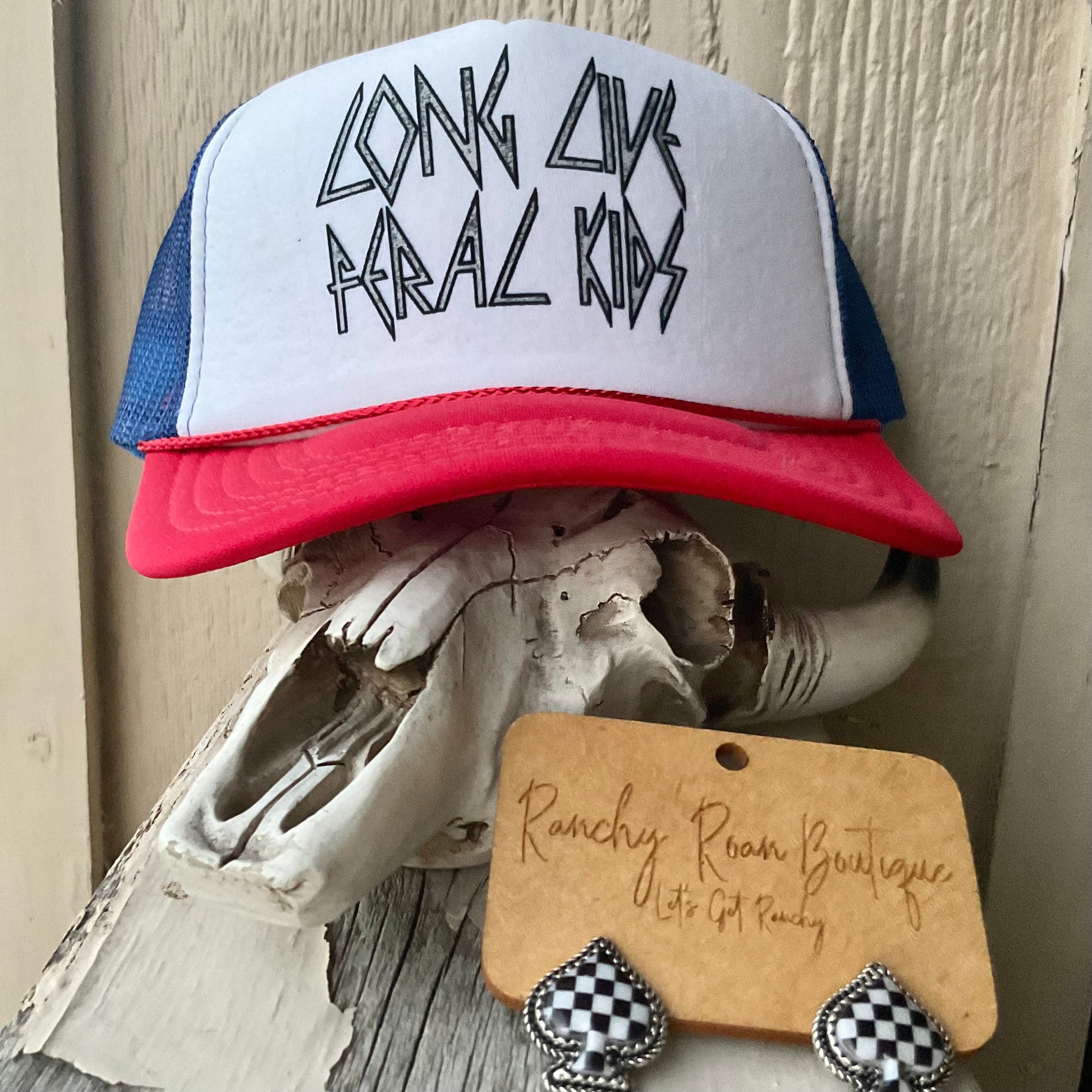 “Long Live Feral Kids” trucker hat displayed on a rustic setting with black-and-white checkered earrings, highlighting its bold, rebellious design.