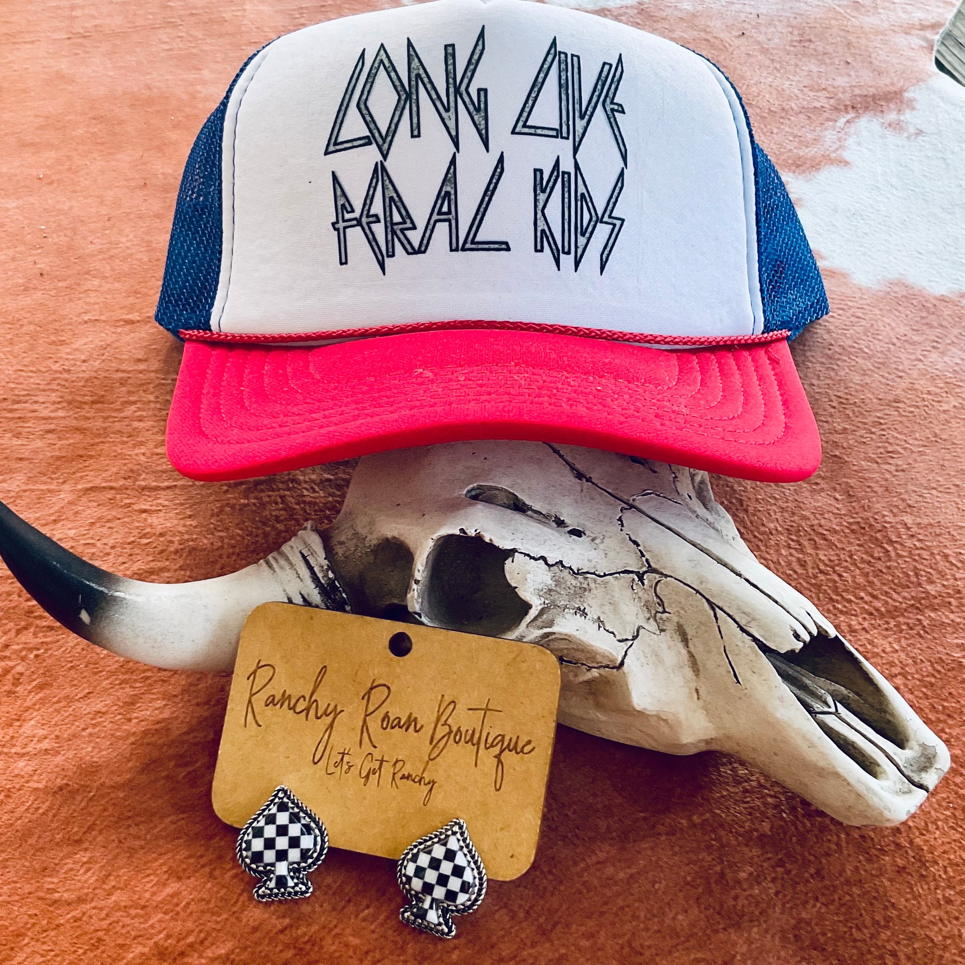 Close-up view of the “Long Live Feral Kids” trucker hat resting on a cow skull, featuring bold text and vibrant red brim with a breathable mesh back.