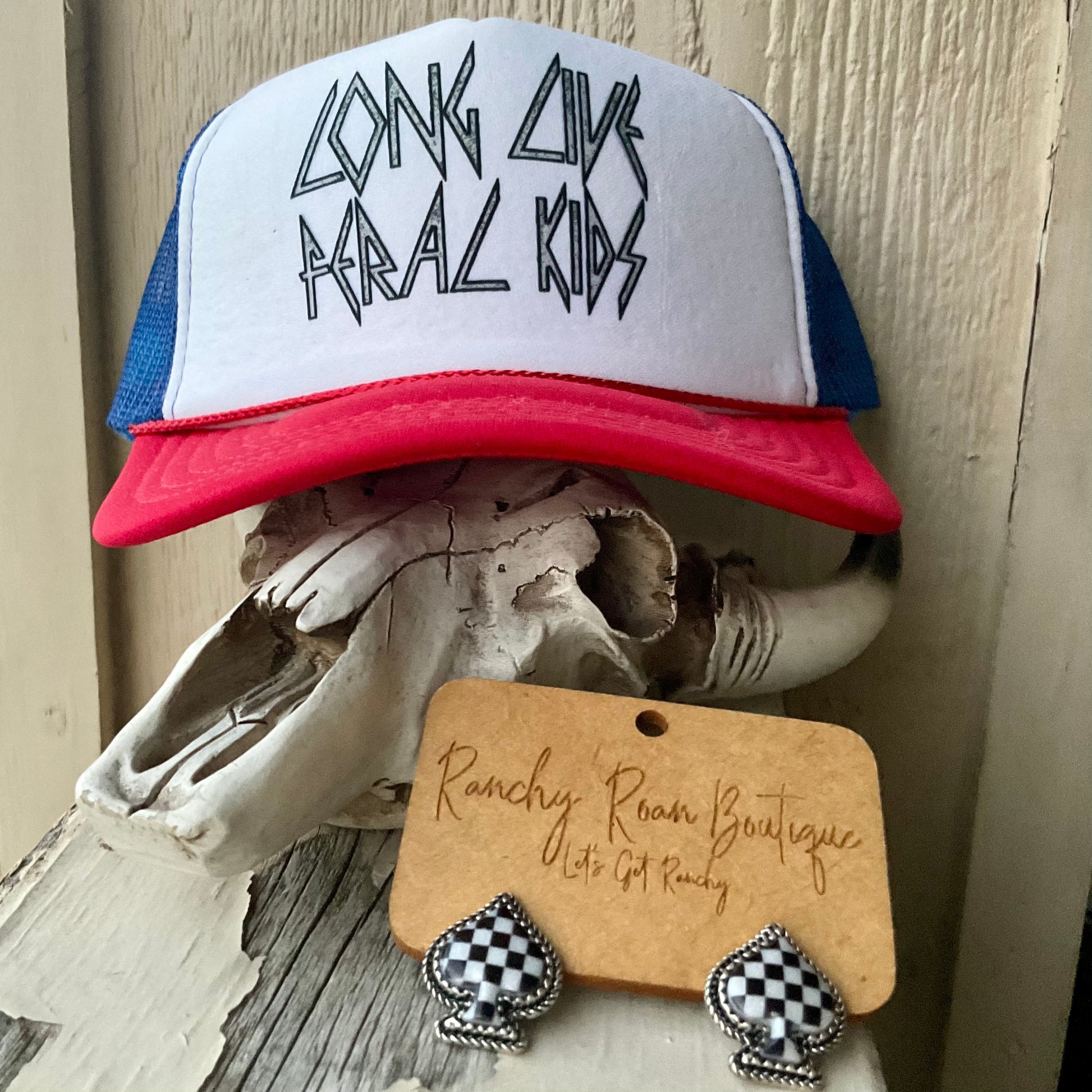 Full display of the “Long Live Feral Kids” trucker hat on a cowhide, showcasing the foam front, red brim, and blue mesh back, styled with Western-themed accessories.