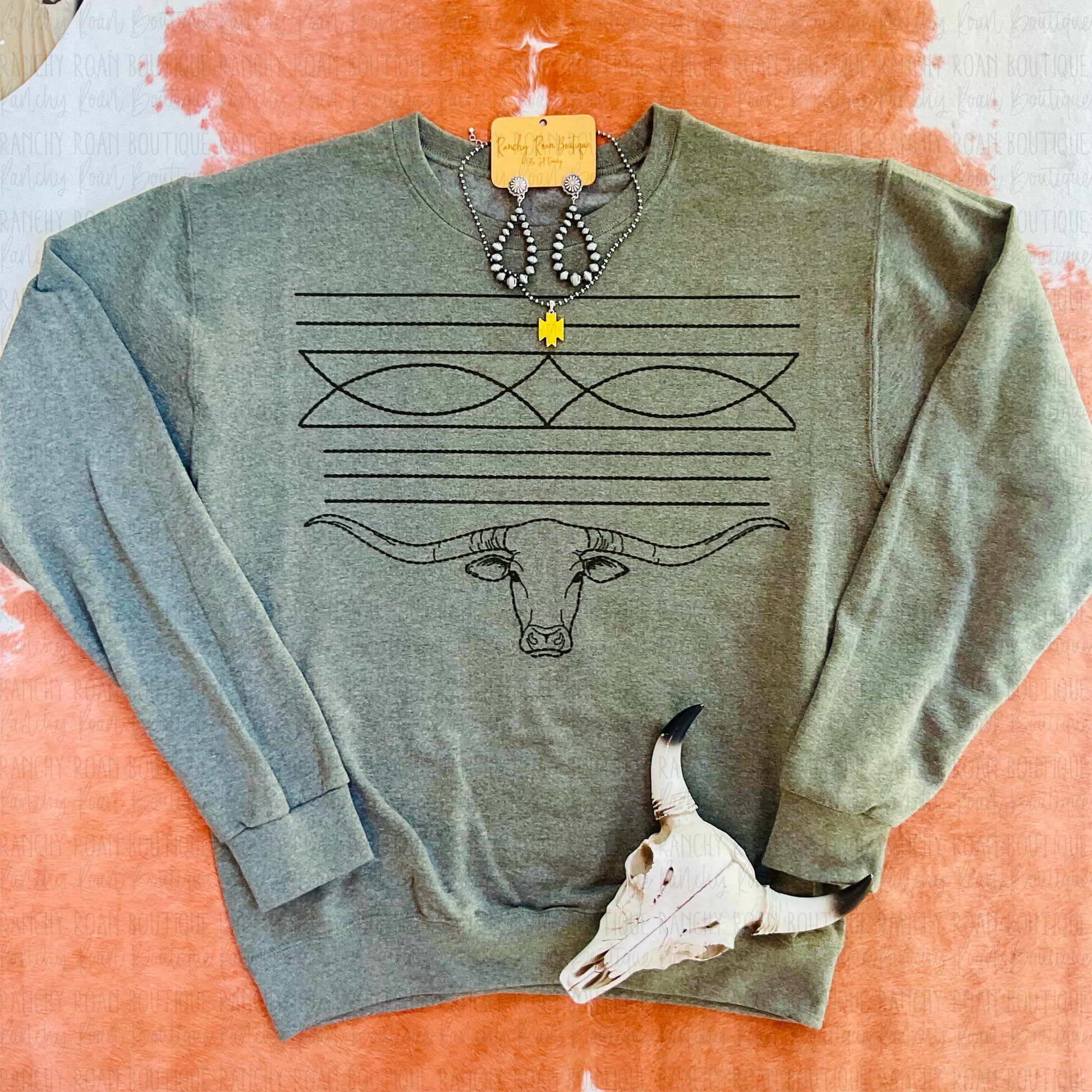 Western Longhorn Boot Stitch Sweatshirt displayed with Western jewelry and a decorative skull on a cowhide background.