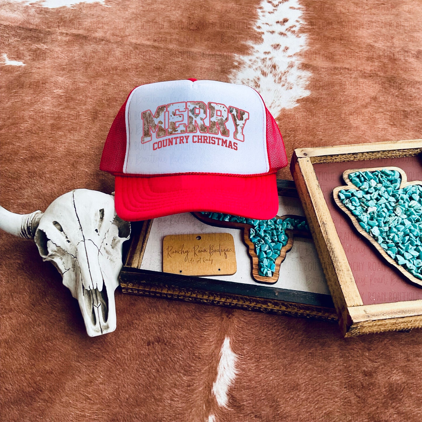 Merry Country Christmas trucker hat featuring a red and white foam front, displayed with turquoise earrings and skull decor on a cowhide background.