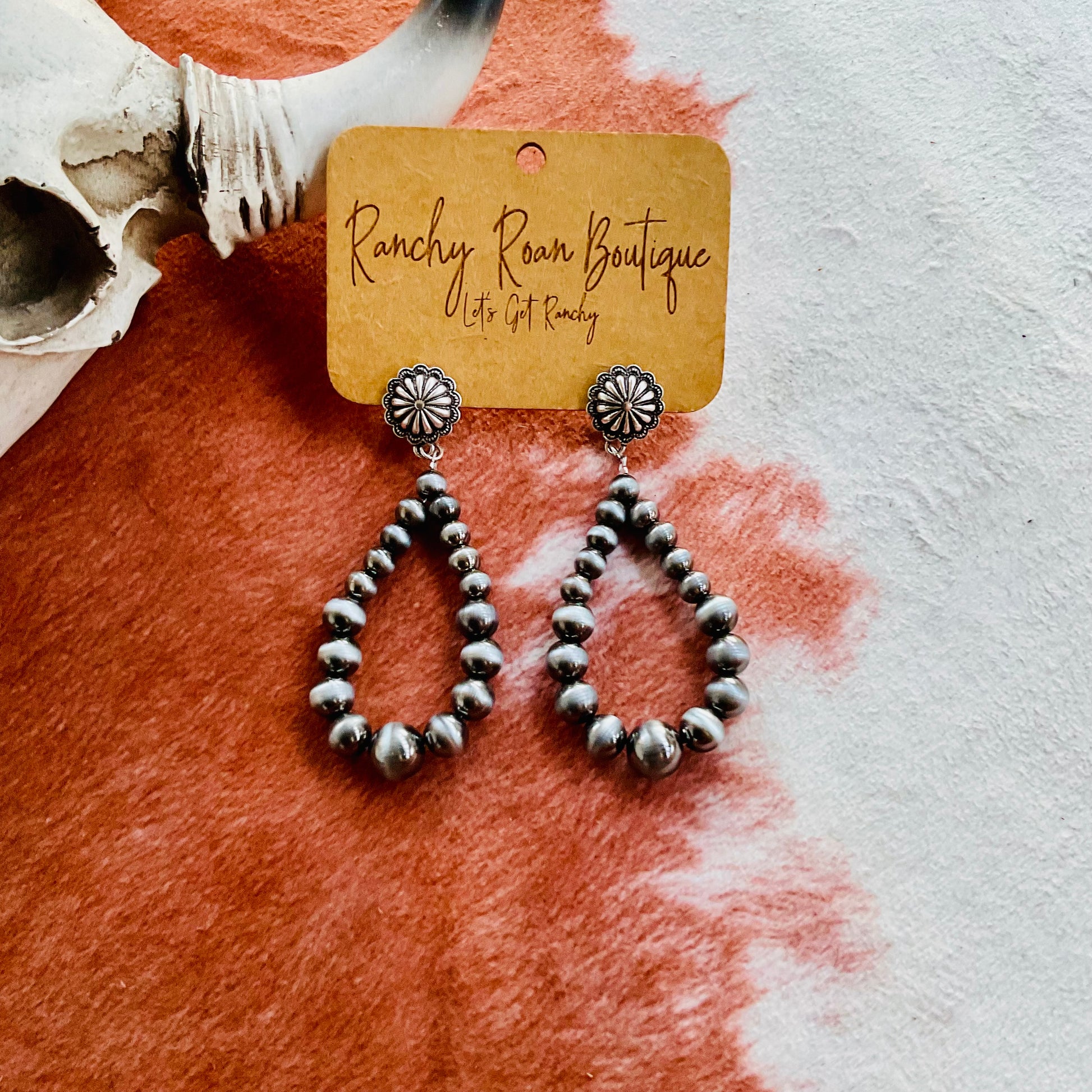 Navajo Style Bead Teardrop Earrings featuring silver-tone beads in a classic Western design, displayed on a Ranchy Roan Boutique card against a cowhide backdrop.