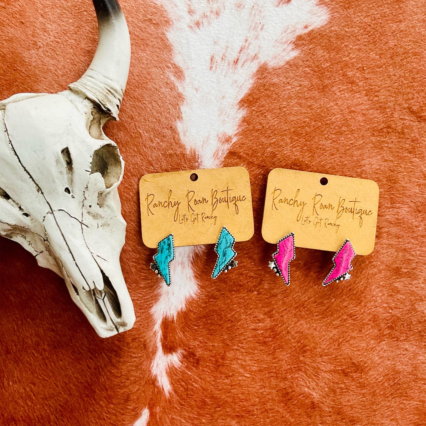 Neon Lightning Bolt Stud Earrings on Cowhide Background: A pair of vibrant neon lightning bolt stud earrings, one in turquoise and the other in hot pink, displayed on a cowhide background with a rustic cow skull. The earrings are mounted on Ranchy Roan Boutique branded cards, highlighting the bold Western-inspired design.