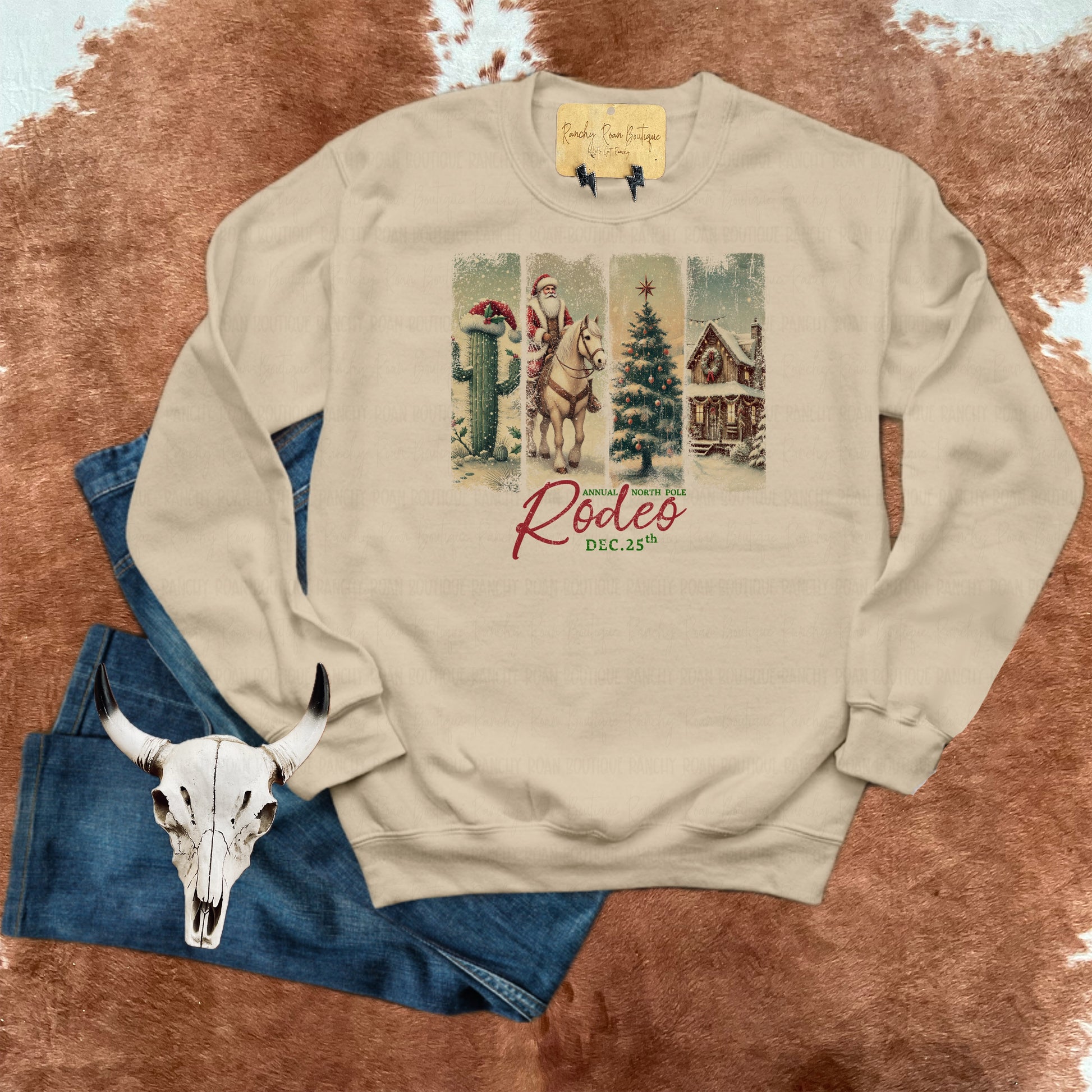 A sandstone sweatshirt featuring a festive North Pole Rodeo Christmas design with Santa on a horse, cacti, a holiday tree, and a rustic cabin, styled with denim jeans and a decorative skull.