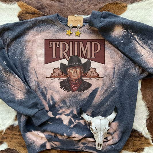 Outlaw Trump Western Sweatshirt with Hand-Bleached Design” - A bold sweatshirt featuring a cowboy-style Trump illustration, displayed on a Western cowhide backdrop with star earrings.