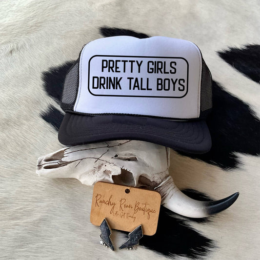 Pretty Girls Drink Tall Boys Western Trucker Hat with bold black text on a white foam front, styled with rustic Western accessories on a cowhide background.