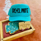 Turquoise foam trucker hat with “Rebel Wife” in bold black lettering and lightning bolt detail, displayed with pink Western earrings and turquoise stones in a wooden tray.
