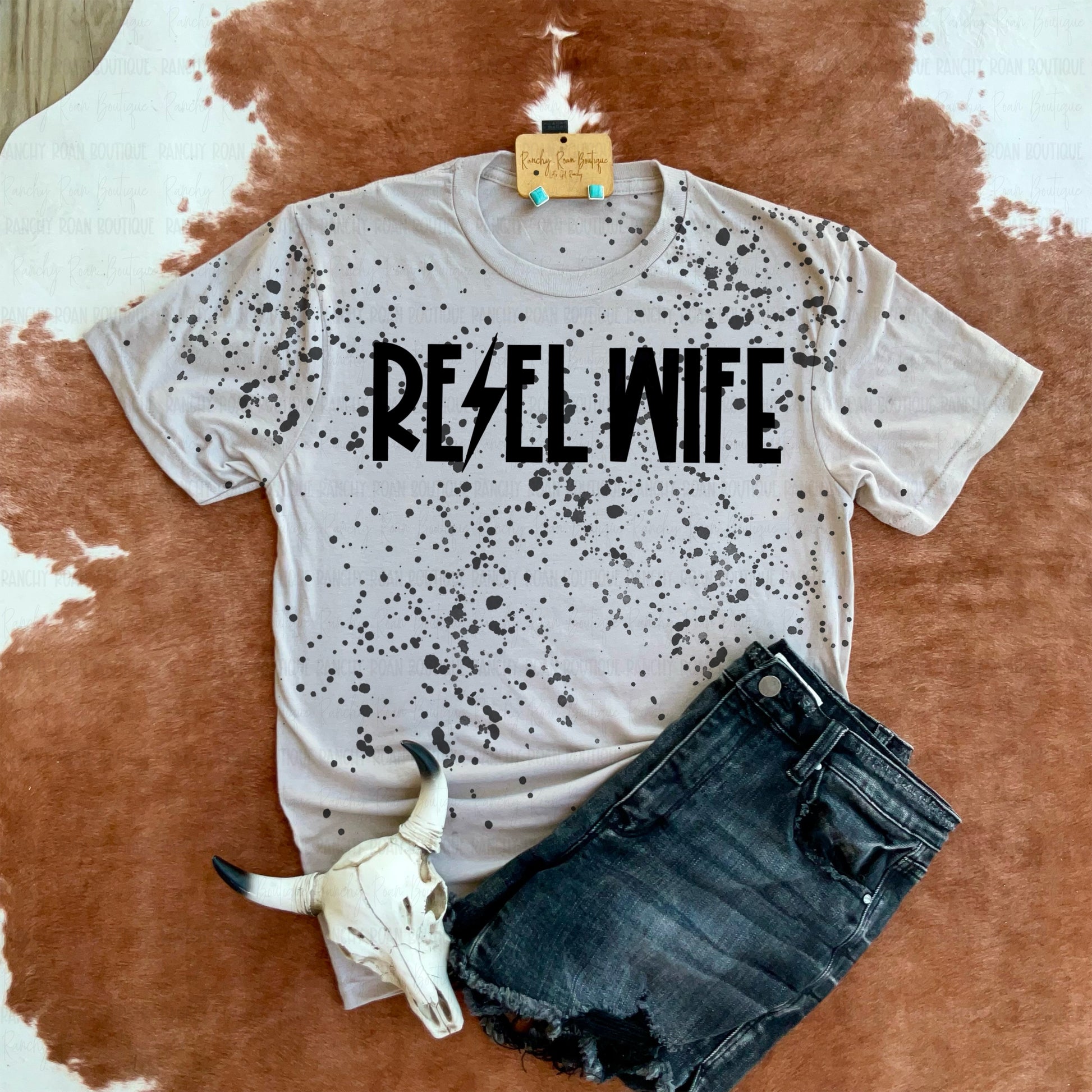 Rebel Wife graphic t-shirt with black splatter pattern and lightning bolt lettering, paired with black denim shorts and displayed on a cowhide background with a decorative skull.