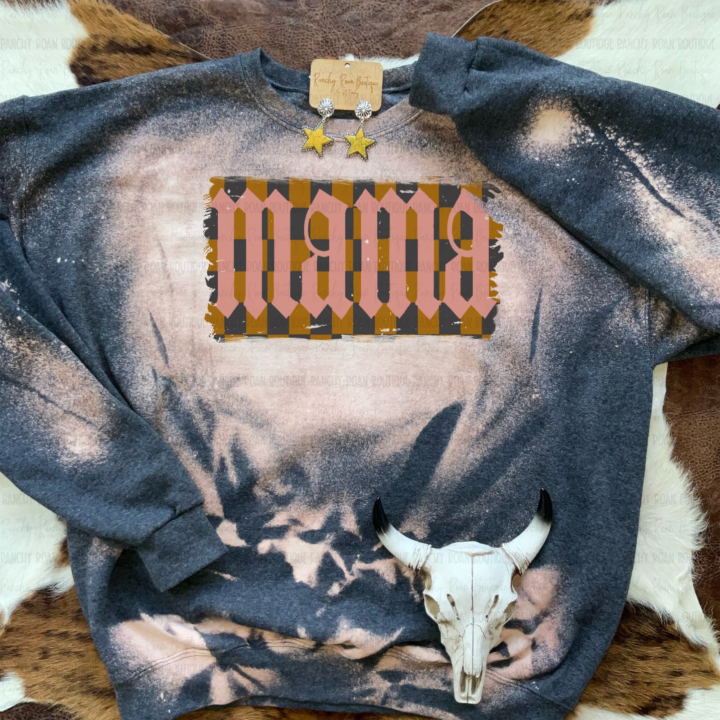 Bleached Western sweatshirt with a retro-inspired “Mama” graphic in warm earth tones, displayed with star earrings and a cow skull on a cowhide background.
