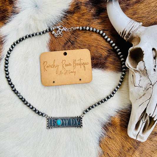 Close-up of the ROWDY bar necklace displayed on a cowhide background, highlighting the burnished silver details and turquoise accents.
