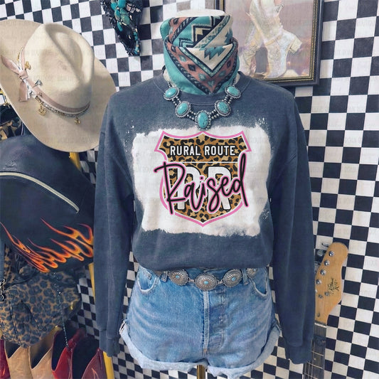 Person wearing a black black heather sweatshirt with 'Rural Route Raised' design, denim shorts, and turquoise jewelry against a checkered wall.