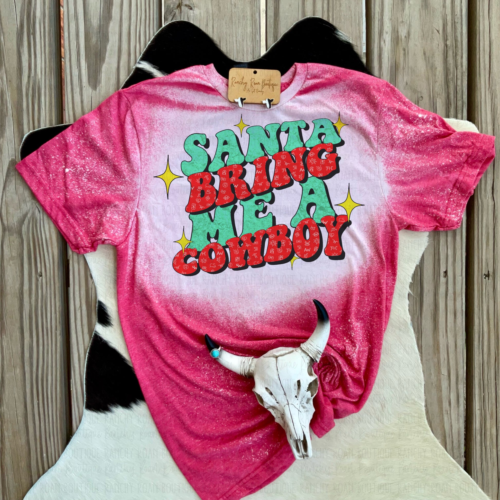 Vintage red bleached “Santa Bring Me a Cowboy” graphic t-shirt displayed with a decorative skull on a wooden background.