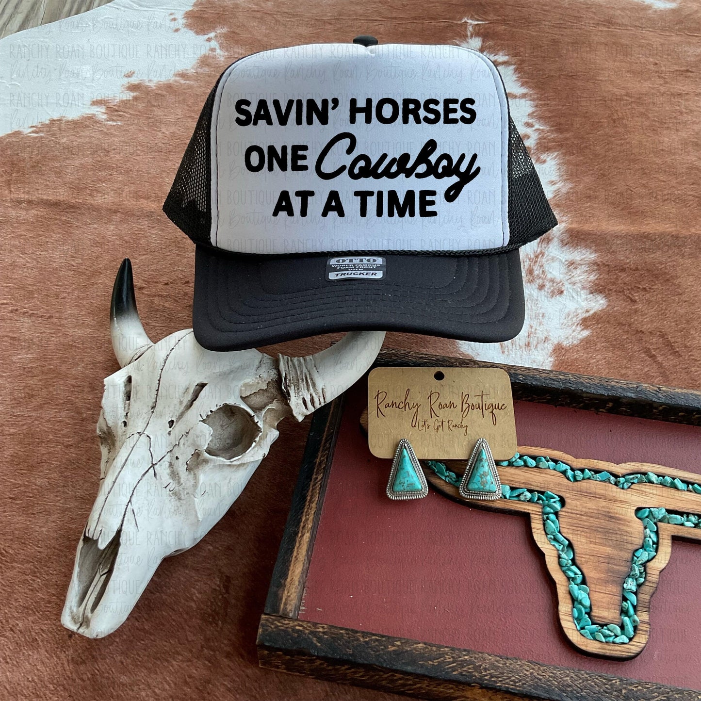 Savin’ Horses One Cowboy at a Time Trucker Hat featuring bold black text on a white foam front, styled with rustic Western accessories.