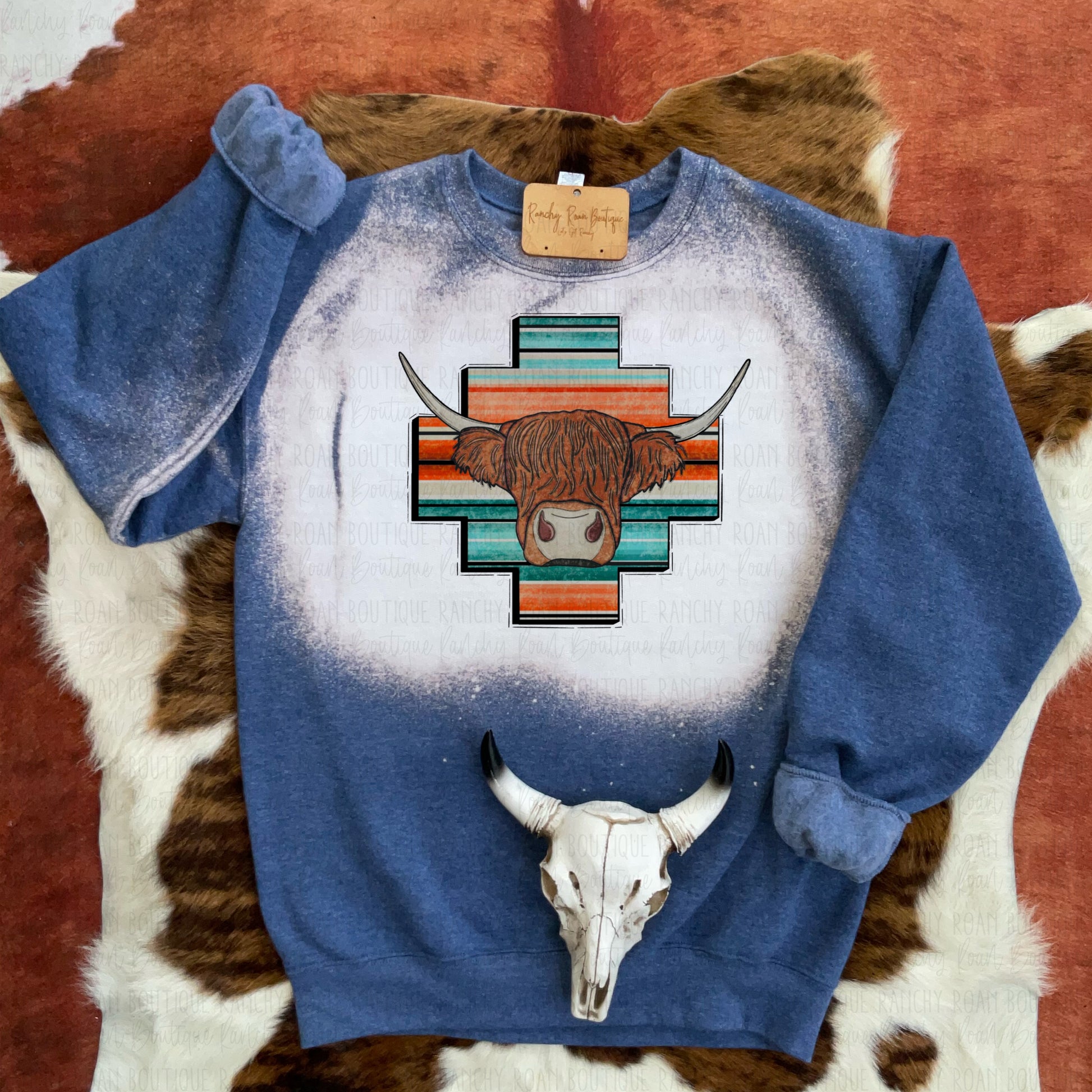 Blue unisex crew sweatshirt with a graphic of a highland cow set against a Serape Aztec design, featuring a unique bleach splatter pattern.