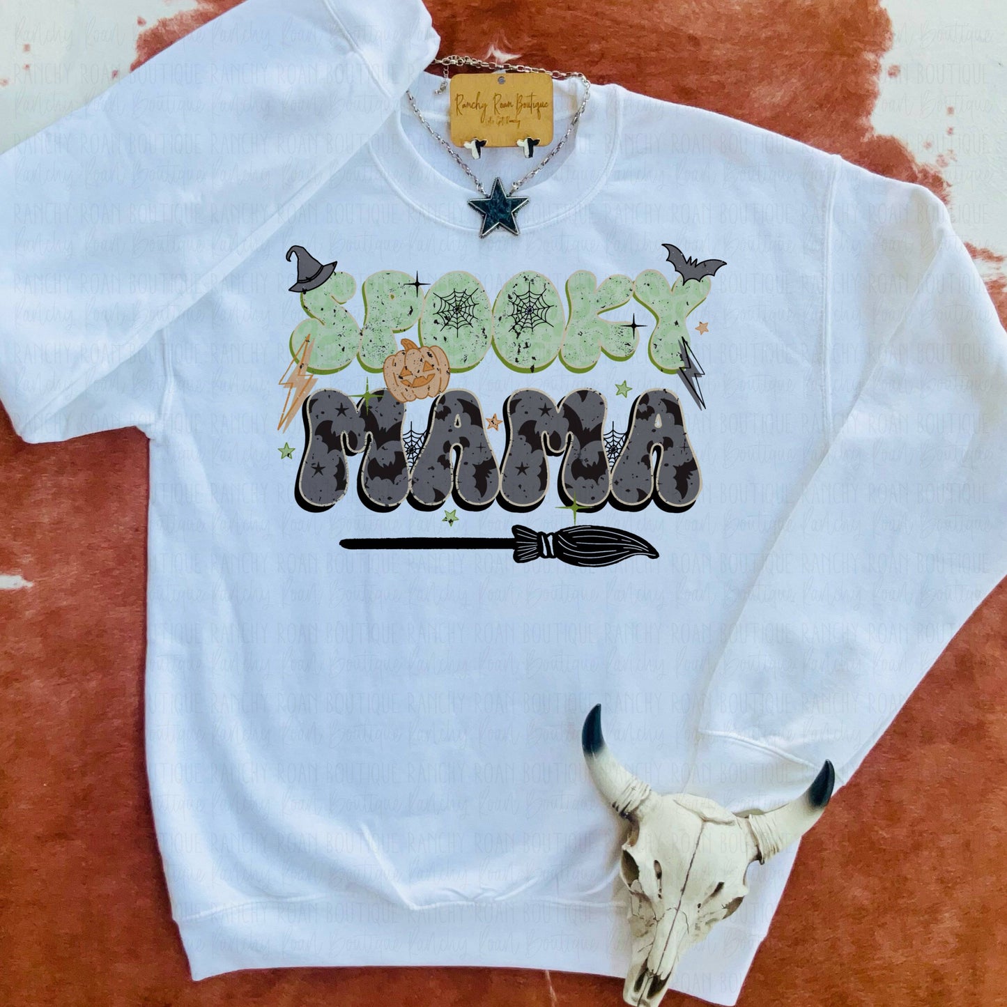 A white sweatshirt from Ranchy Roan Boutique featuring the text “Spooky Mama” in bold letters with leopard print and Halloween-themed elements like witch hats and a broomstick. Styled with turquoise star jewelry and a cow skull accessory, emphasizing the Western aesthetic.