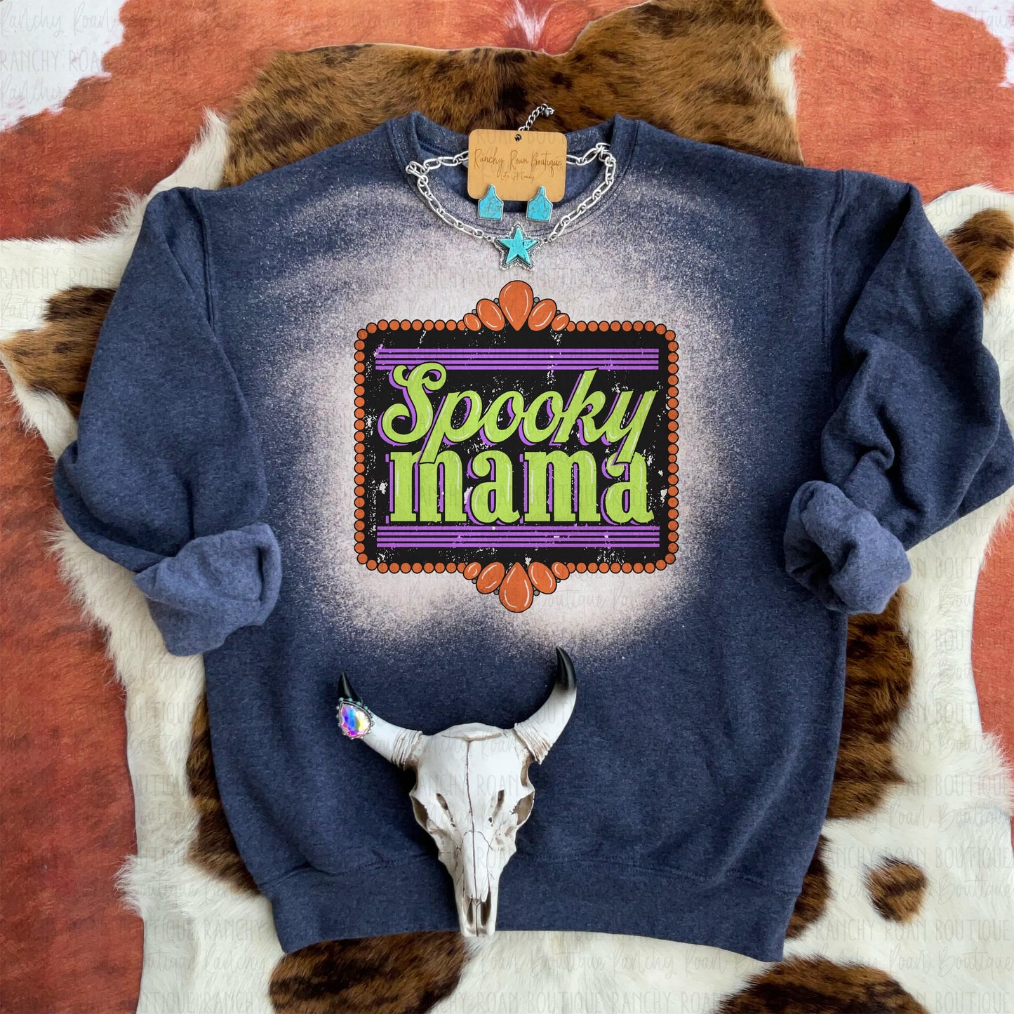 A bleached sweatshirt from Ranchy Roan Boutique featuring a vibrant sublimation print with the text “Spooky Mama” in bold, festive colors. The sweatshirt has a unique bleached pattern in shades of dark blue and cream, creating a one-of-a-kind look. Styled with a turquoise star necklace and a cow skull accessory, enhancing the Western aesthetic. The background includes a cowhide texture, further emphasizing the rustic, Western theme.