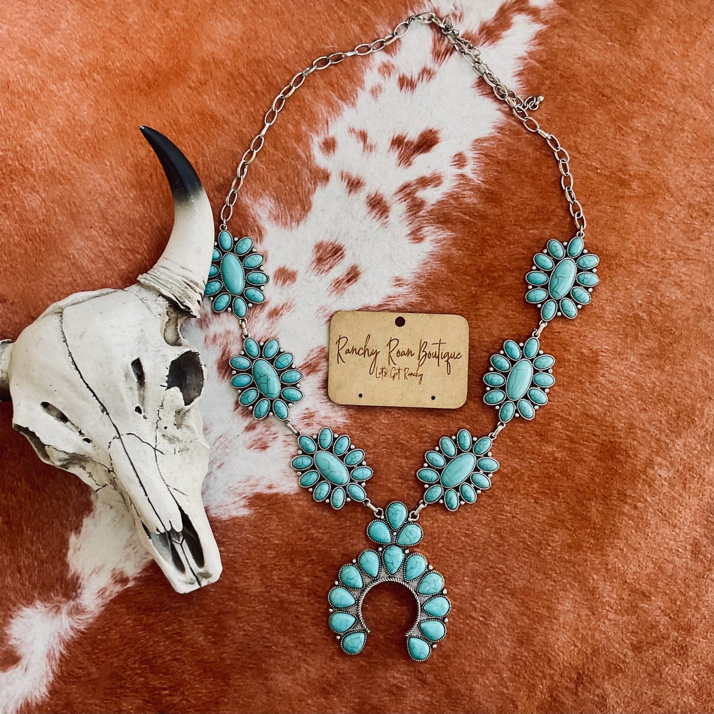 Turquoise stone squash blossom necklace in burnished silver-tone, displayed with Western cow skull decor against a cowhide background.