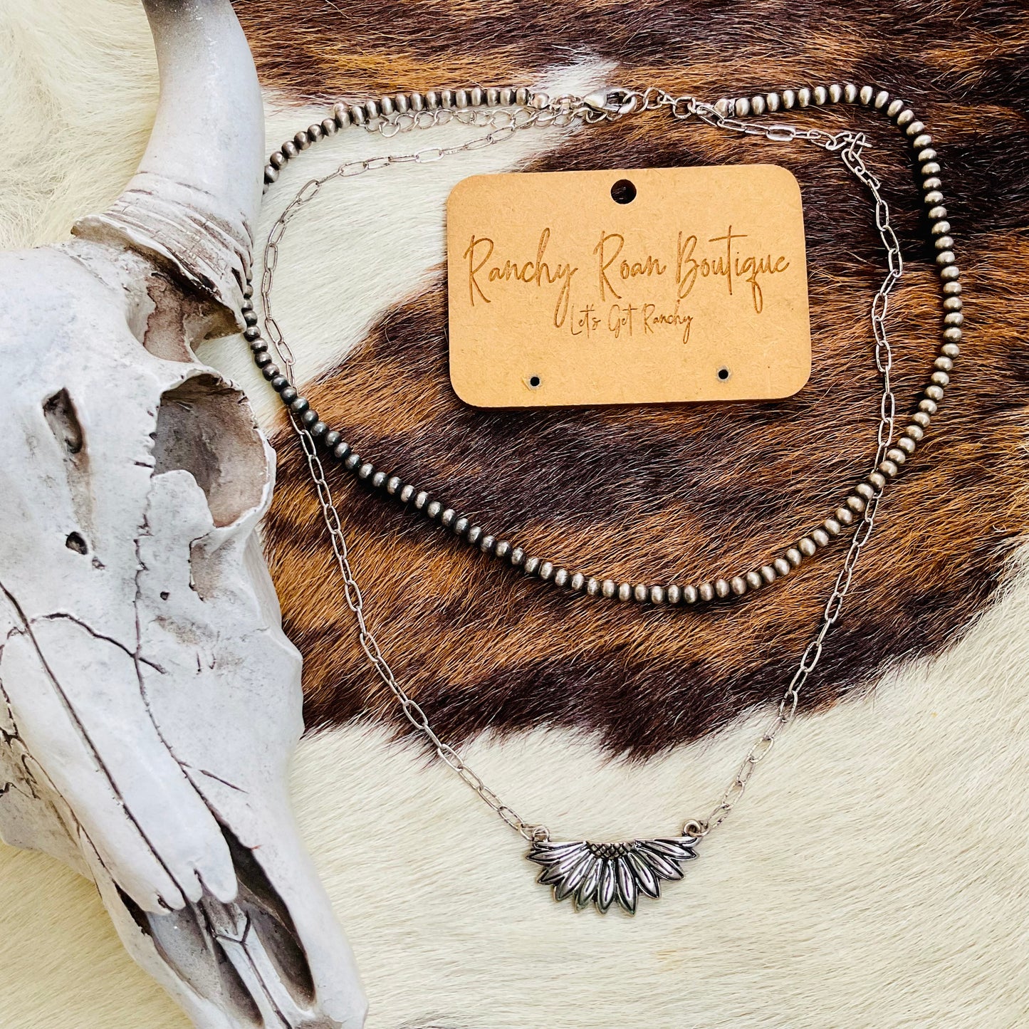 Close-up view of a Sunflower Casting Two-Row Necklace displayed on a cowhide background, showcasing its burnished silver design and dual-layer chains.