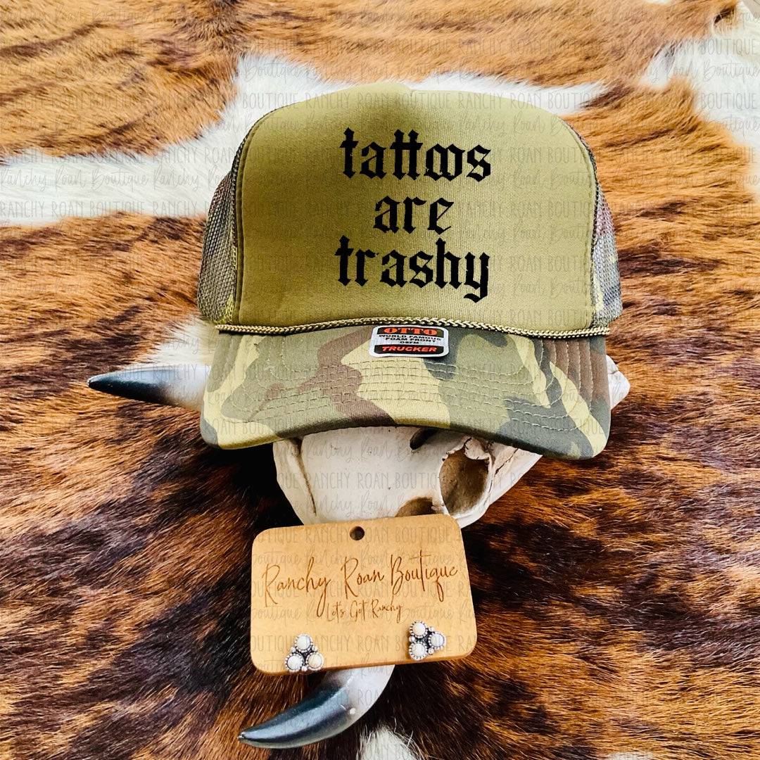 Tattoos Are Trashy Camo Trucker Hat featuring bold black text on an olive green background with camo accents, styled on a rustic Western backdrop