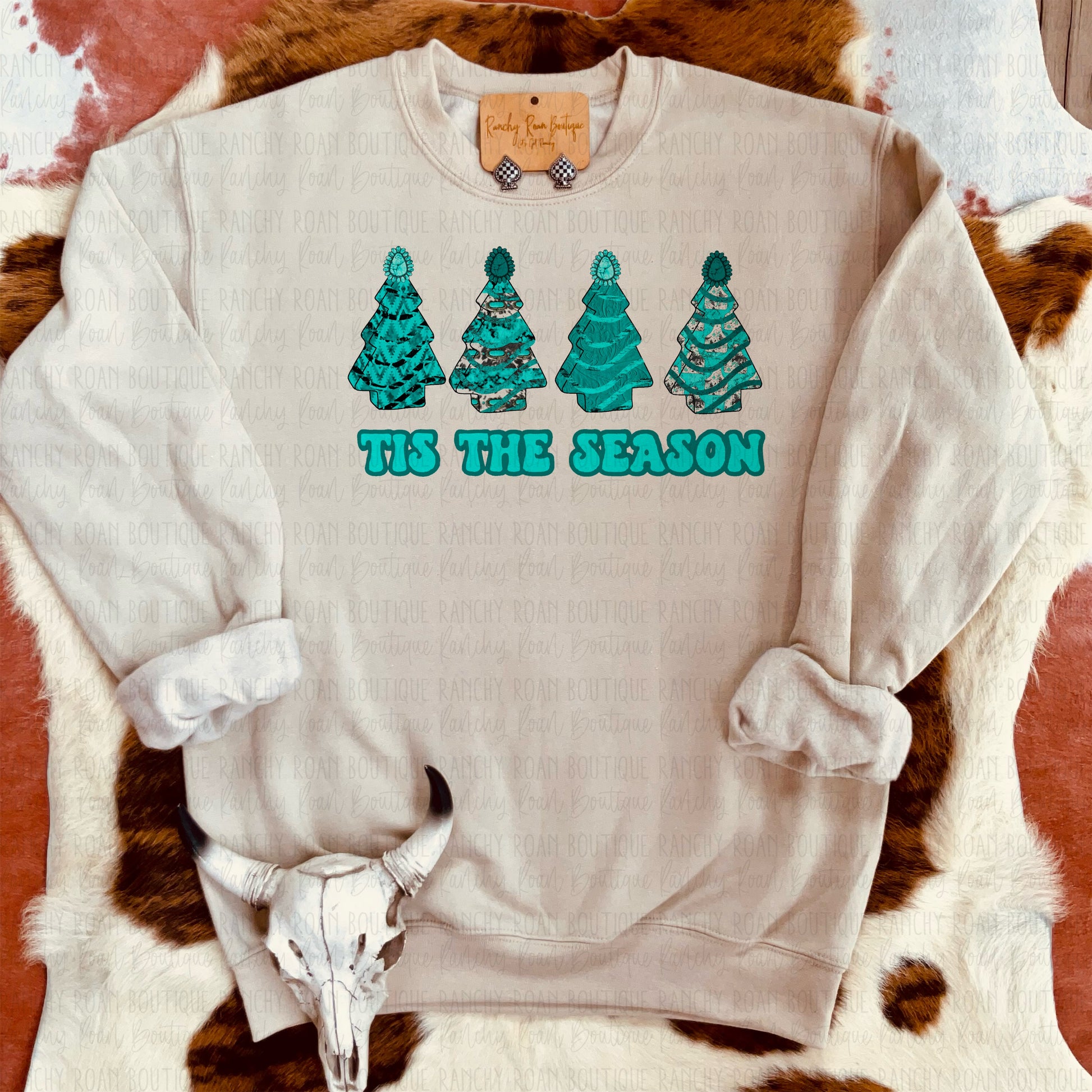 Sandstone sweatshirt with turquoise Christmas tree designs and “Tis the Season” text, styled against a Western backdrop.