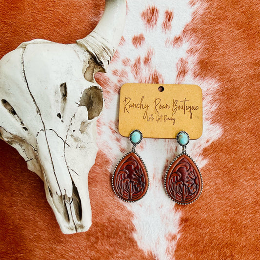 Tooled Leather and Turquoise Teardrop Earrings with burnished silver edge, displayed on a rustic background with a cow skull.