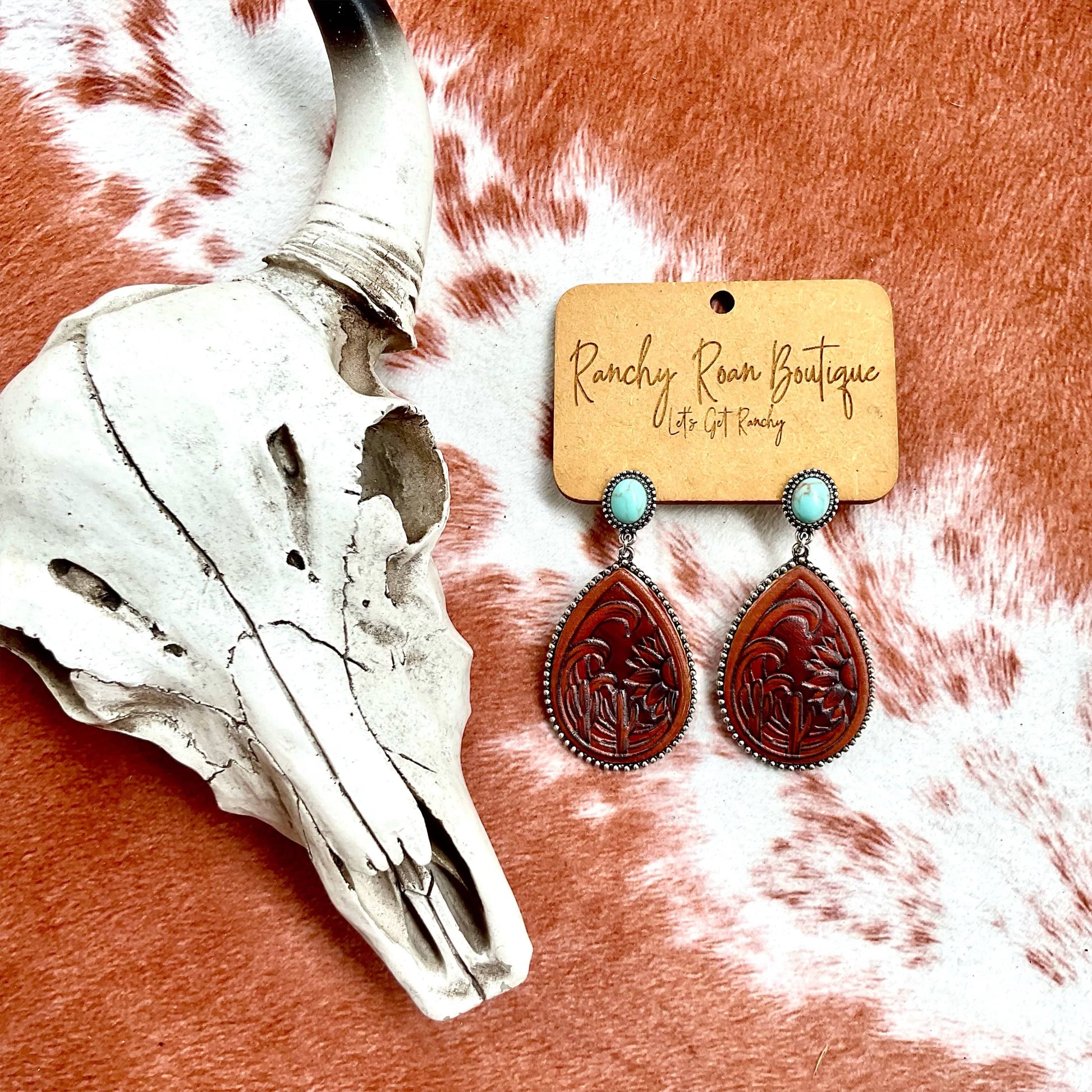 Tooled Leather and Turquoise Teardrop Earrings with burnished silver edge, displayed on a rustic background with a cow skull.