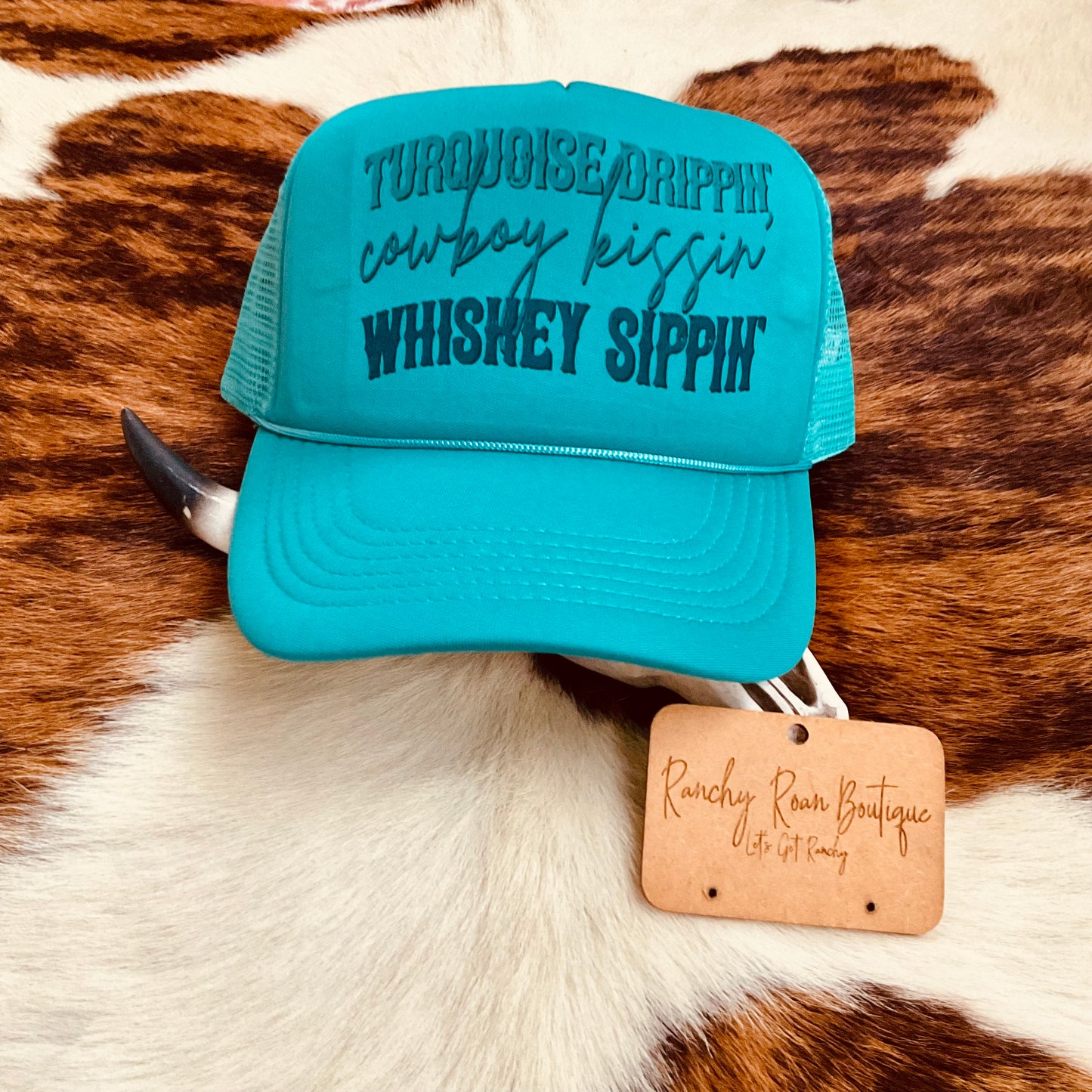 Turquoise trucker hat with ‘Turquoise Drippin’, Cowboy Kissin’, Whiskey Sippin’’ slogan styled with a cow skull and ranchy branding