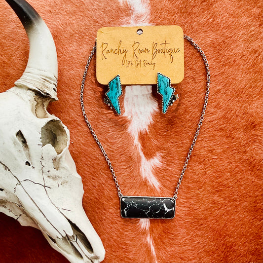 A set featuring turquoise lightning bolt earrings and a black stone bar necklace displayed on a Ranchy Roan Boutique card, set against a cowhide background with a decorative cow skull
