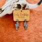  A pair of turquoise-accented checkered lightning bolt earrings displayed on a Ranchy Roan Boutique card against a cowhide background with a decorative cow skull.