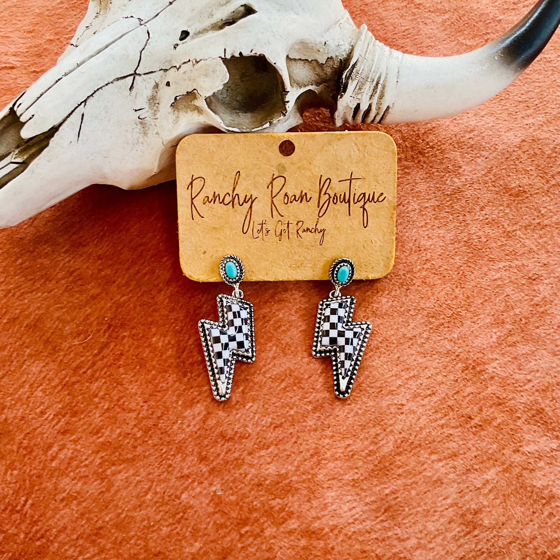  A pair of turquoise-accented checkered lightning bolt earrings displayed on a Ranchy Roan Boutique card against a cowhide background with a decorative cow skull.