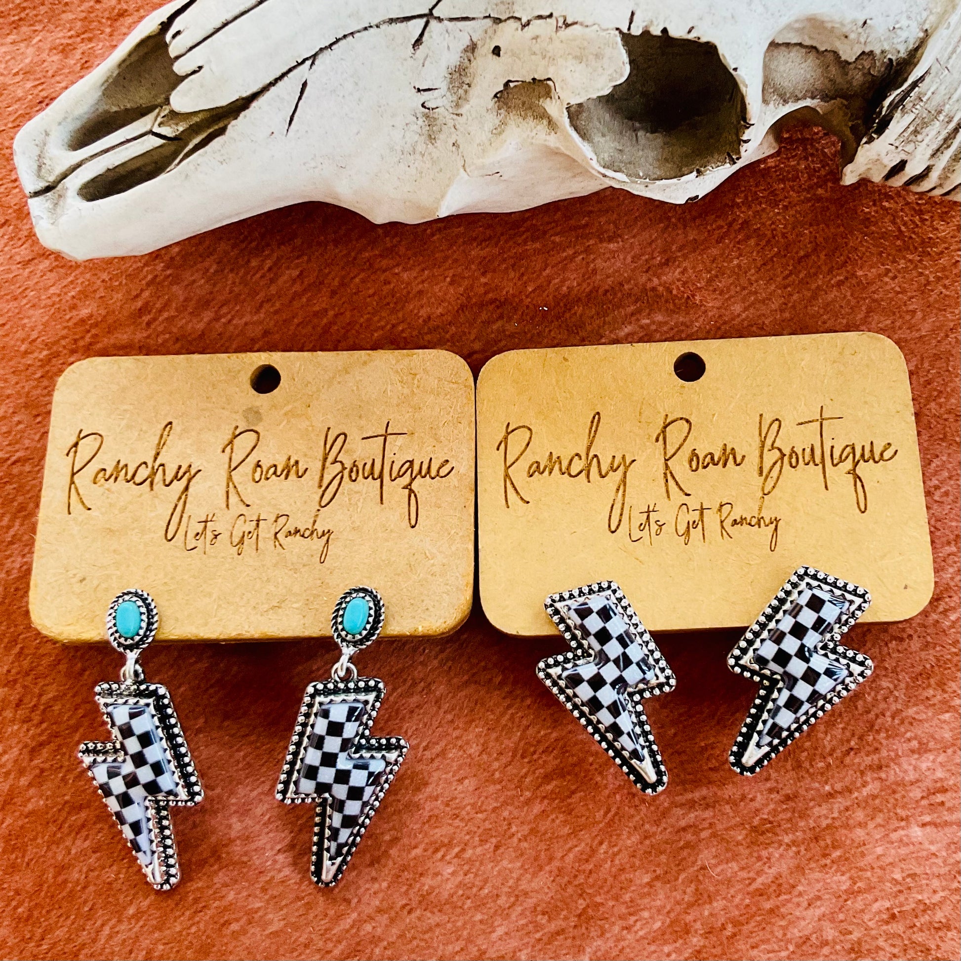 A pair of turquoise-accented checkered lightning bolt earrings and regular checkered lightning bolt earrings displayed on Ranchy Roan Boutique cards against a cowhide background with a decorative cow skull.
