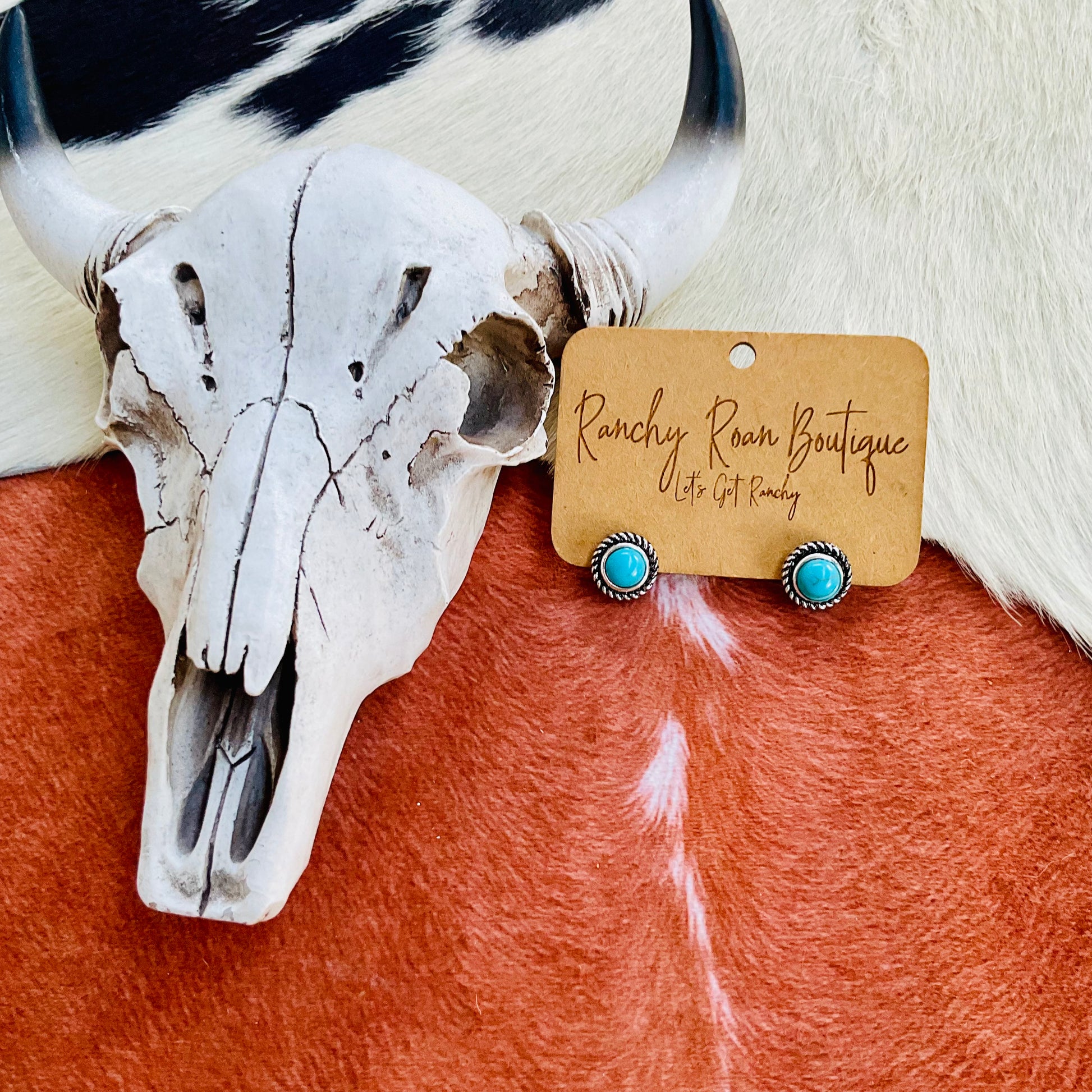  A pair of turquoise stud earrings with silver detailing, displayed on a Ranchy Roan Boutique card against a cowhide backdrop with a decorative cow skull.