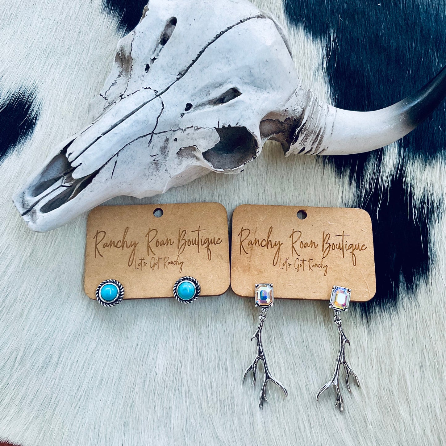  A pair of turquoise stud earrings and crystal antler dangle earrings displayed on Ranchy Roan Boutique cards, set against a cowhide background with a decorative cow skull.
