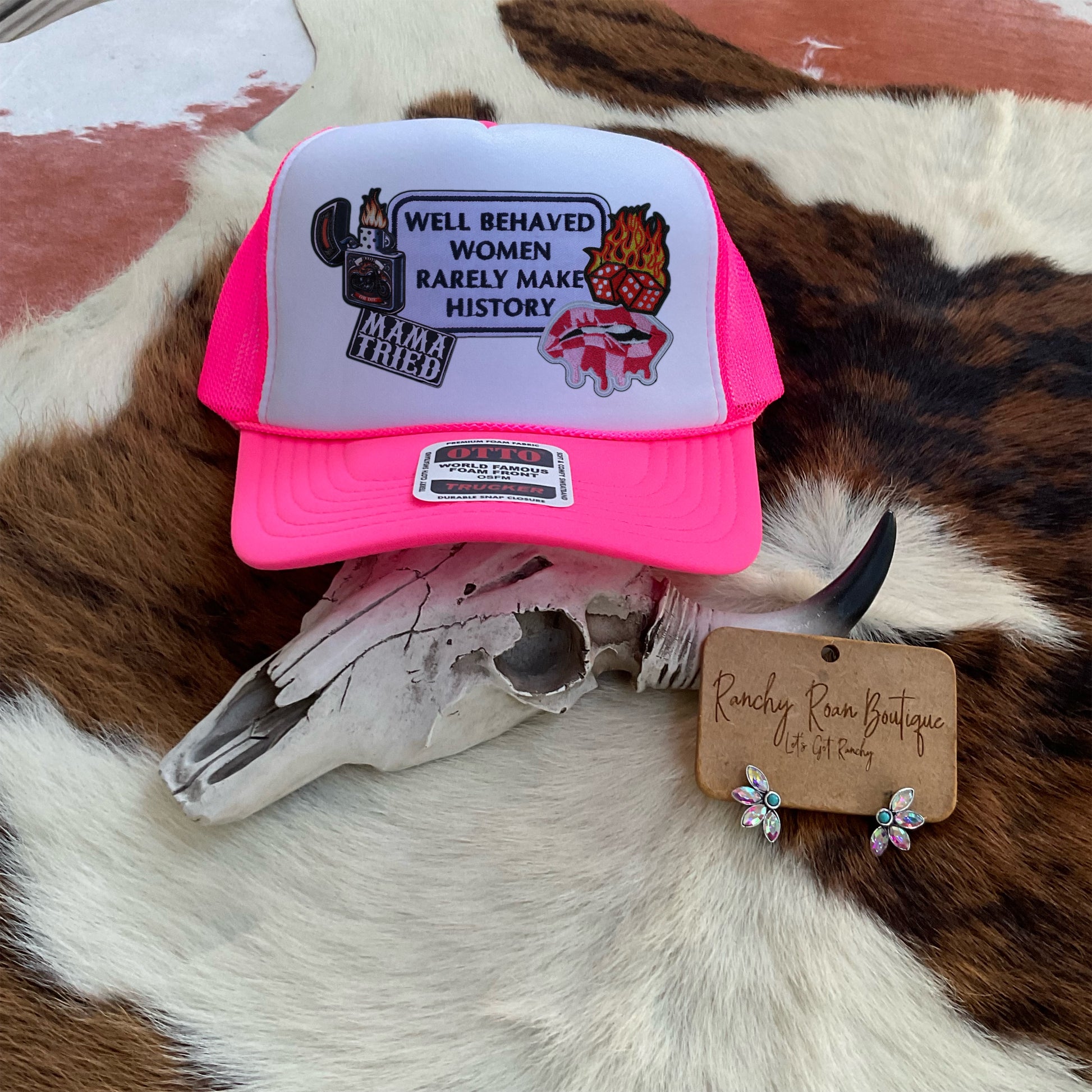  Hot pink trucker hat with faux-patch graphics, including flames, dice, and bold retro text, displayed on a Western cowhide backdrop.
