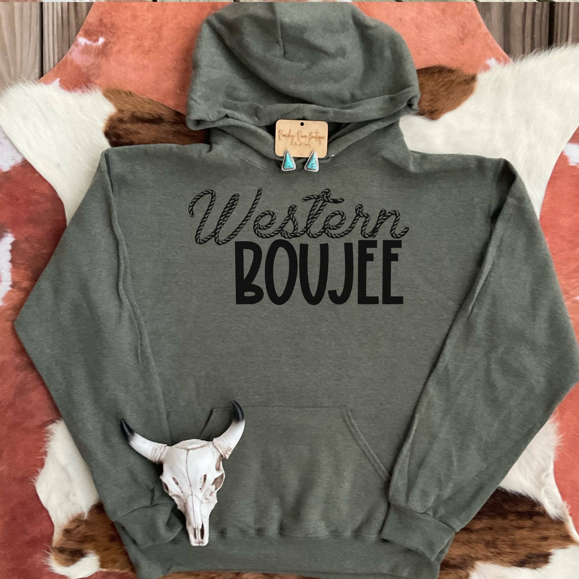 A heather military green hoodie from Ranchy Roan Boutique featuring a bold sublimation print with the text “Western Boujee” in a stylish font. The hoodie is displayed on a cowhide background, styled with turquoise earrings and a cow skull accessory, enhancing the Western aesthetic.