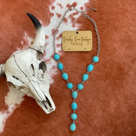  Burnished silver Y-shaped necklace with turquoise cabochon stones, displayed with Western cow skull decor against a cowhide background.
