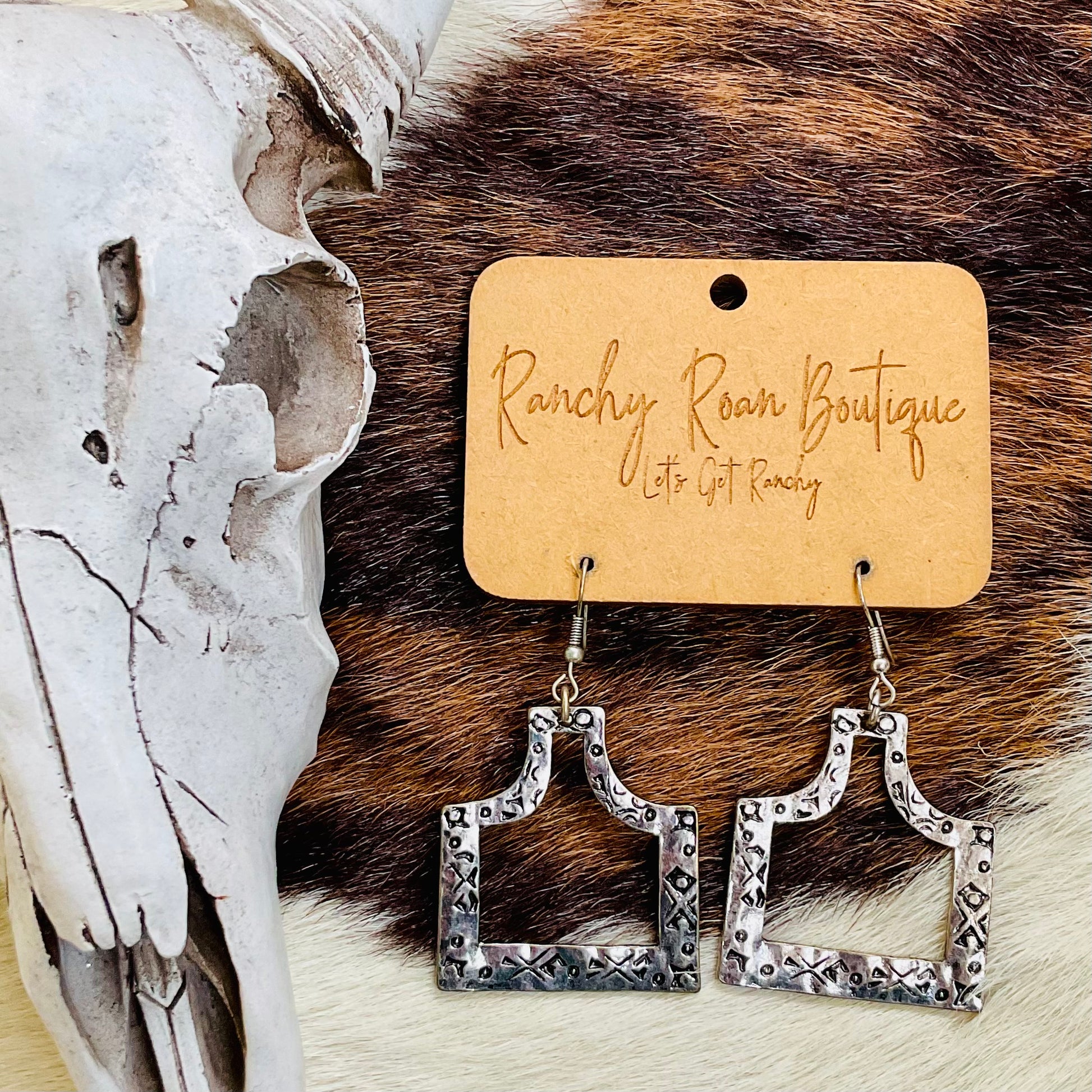 Western Cattle Tag Cutout Dangle Earrings displayed on a ranchy background with cowhide and skull decor.